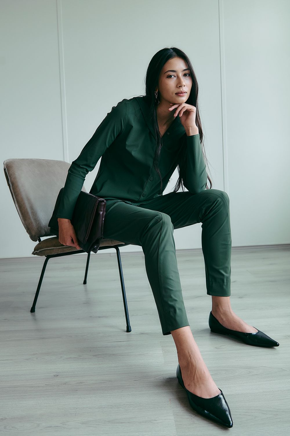 A series of fashion poses showing a person in an emerald green pantsuit and black flats sitting on a modern chair.