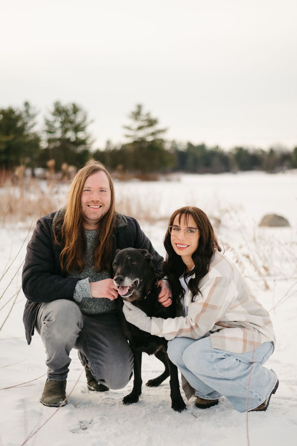 Two people pose with a black dog in snowy winter field during outdoor photo session.