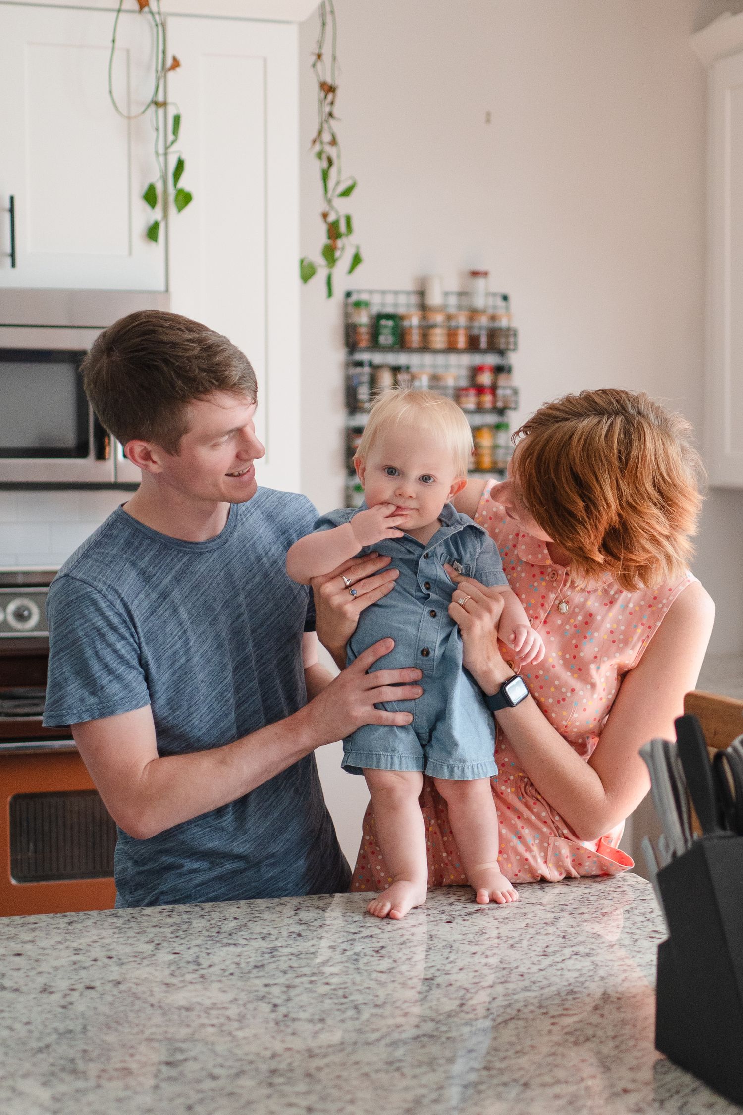 Family moment in modern kitchen as baby in denim stands on counter between caregivers.
