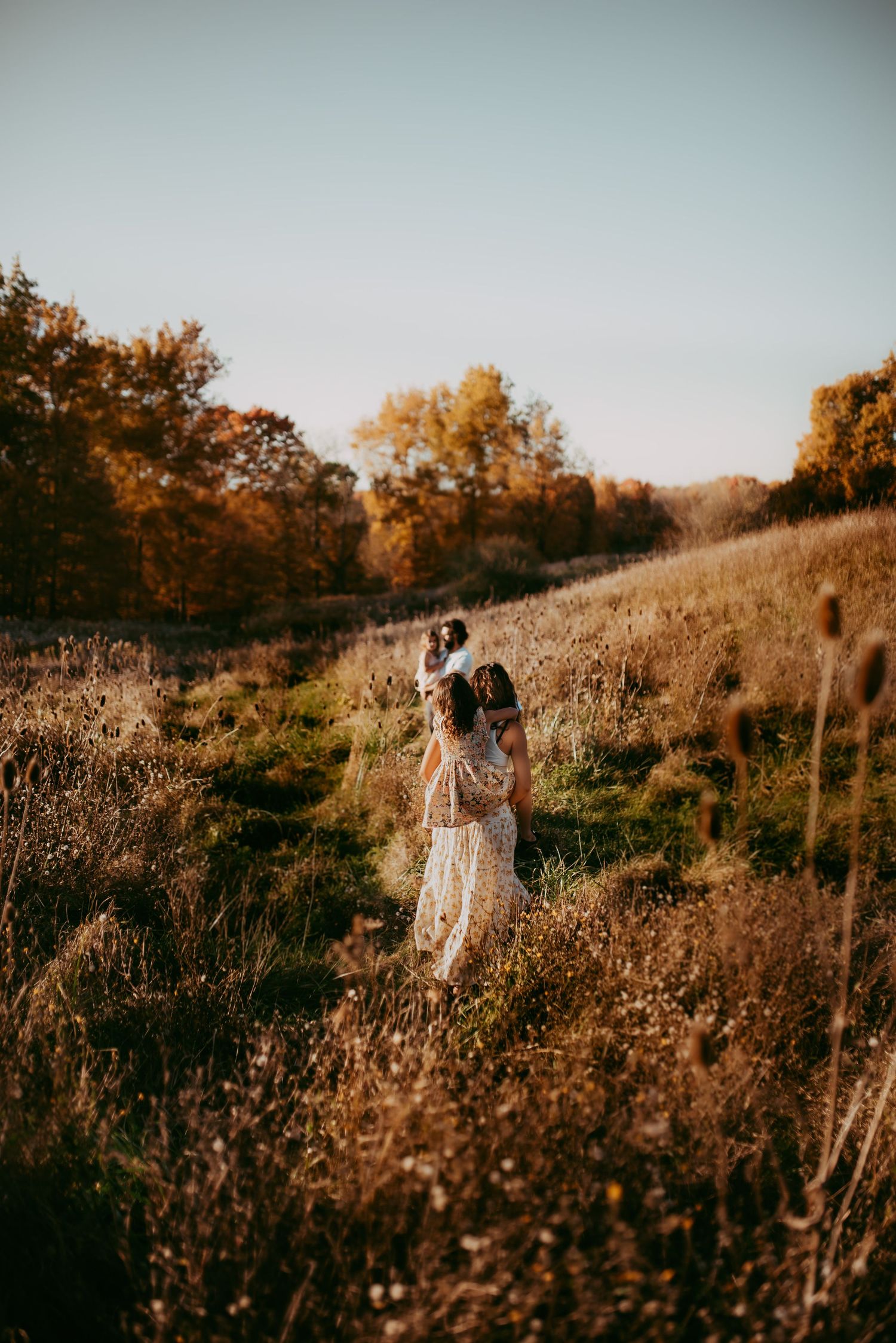 A dreamy fall meadow scene at golden hour shows a figure in a white dress walking through tall dried grasses carrying a child