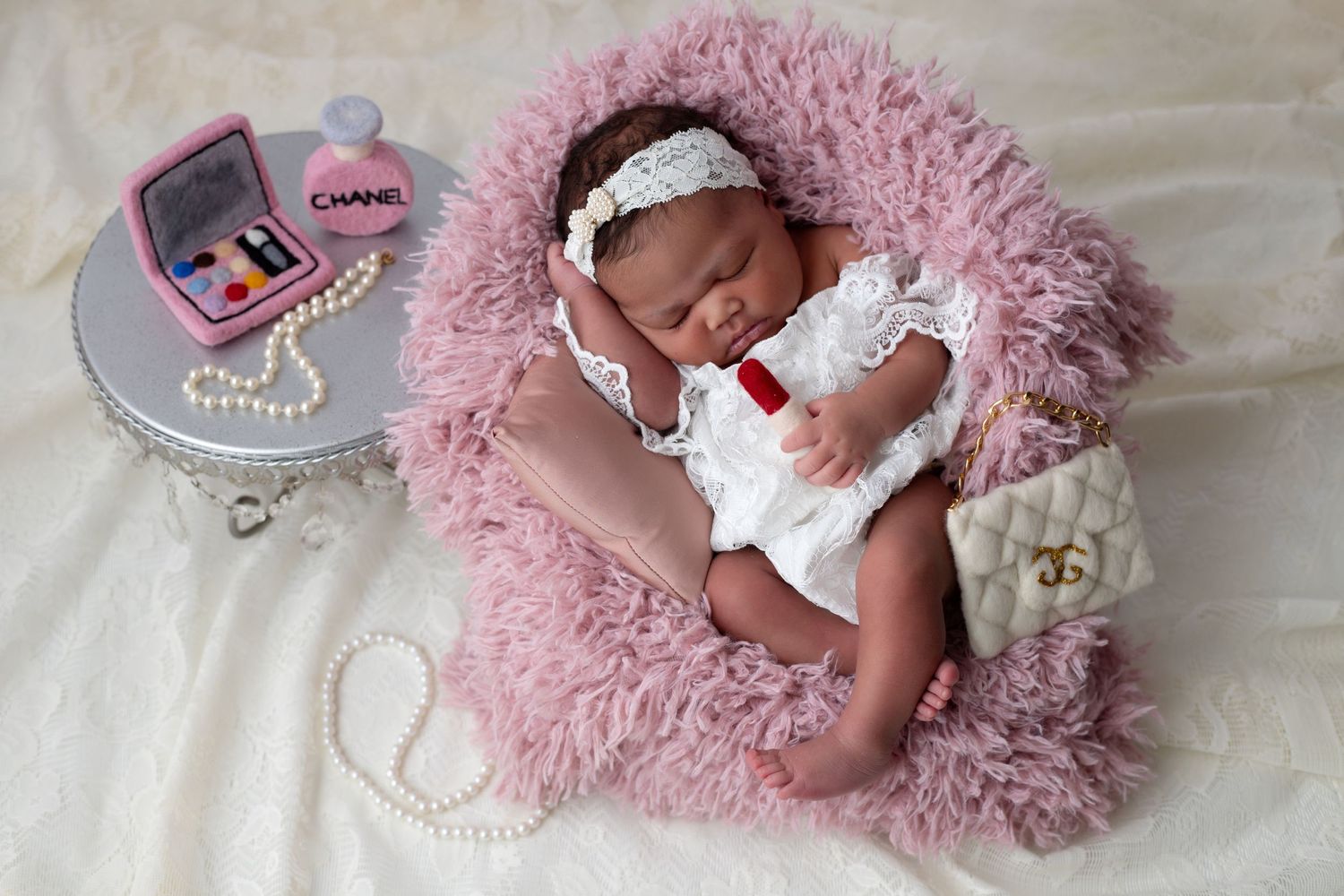 Newborn baby girl peacefully sleeping on a soft pink fur chair surrounded by Chanel make-up, purse and accessories for newborn photoshoot.
