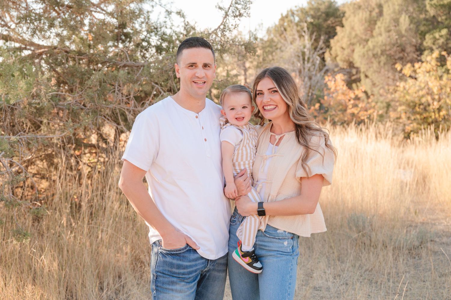 Small family poses together in golden sunlit field during sunset portrait session.