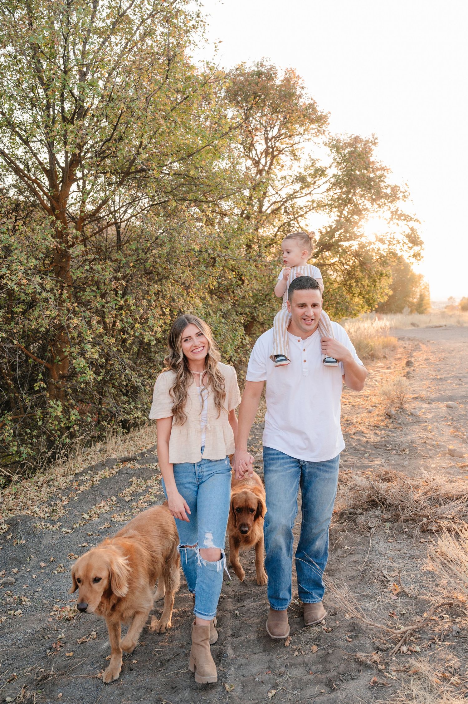 Couple walks their two golden retrievers along trail during sunset with trees in background.