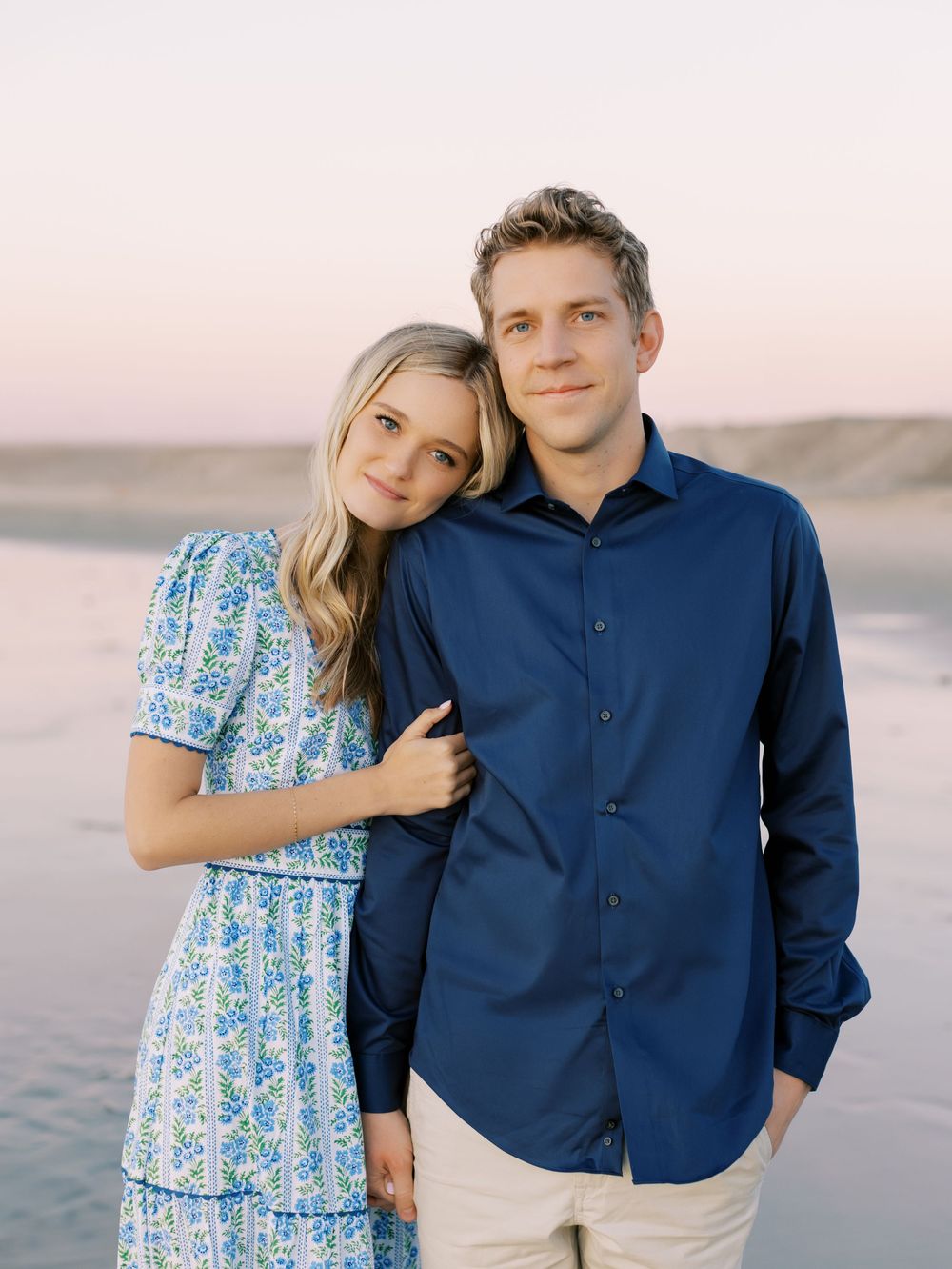 A couple stands together on a beach at sunset wearing coordinated blue and white outfits.