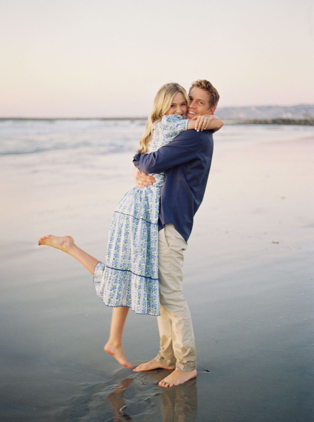 Two people embrace and dance barefoot on a wet sandy beach during sunset.