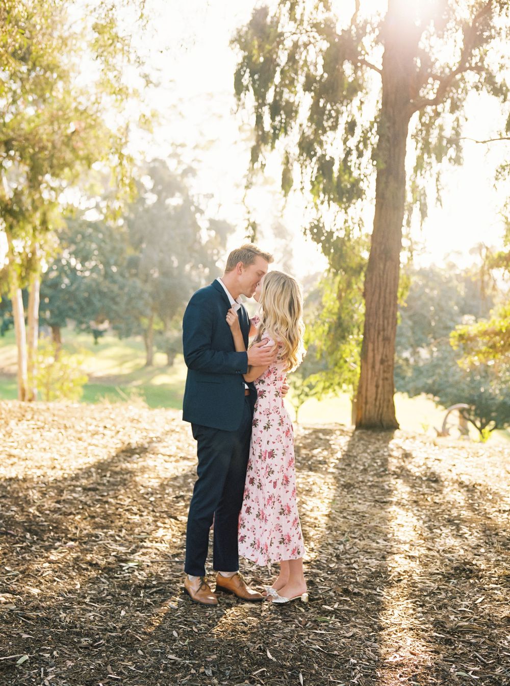 A couple shares a romantic moment at sunset under willow trees in a dreamy outdoor setting.