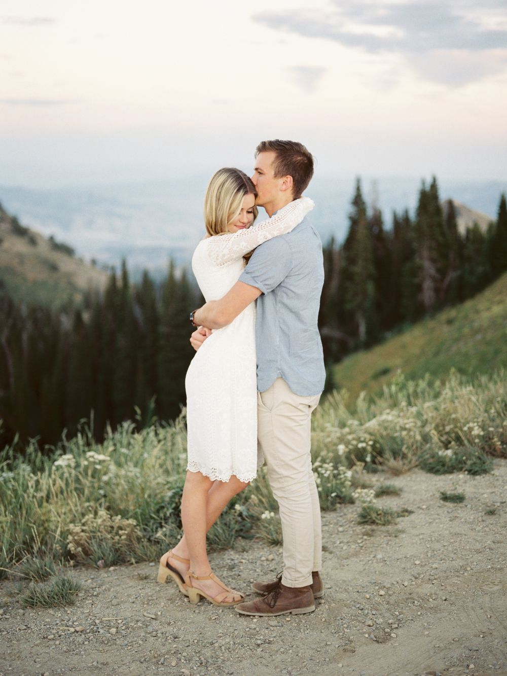 A romantic couple embraces on a mountaintop trail with evergreen trees and mountain views in the background.