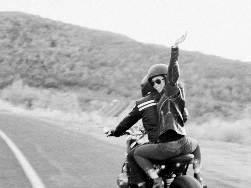 A person rides a motorcycle on an open road with arms raised in celebration.