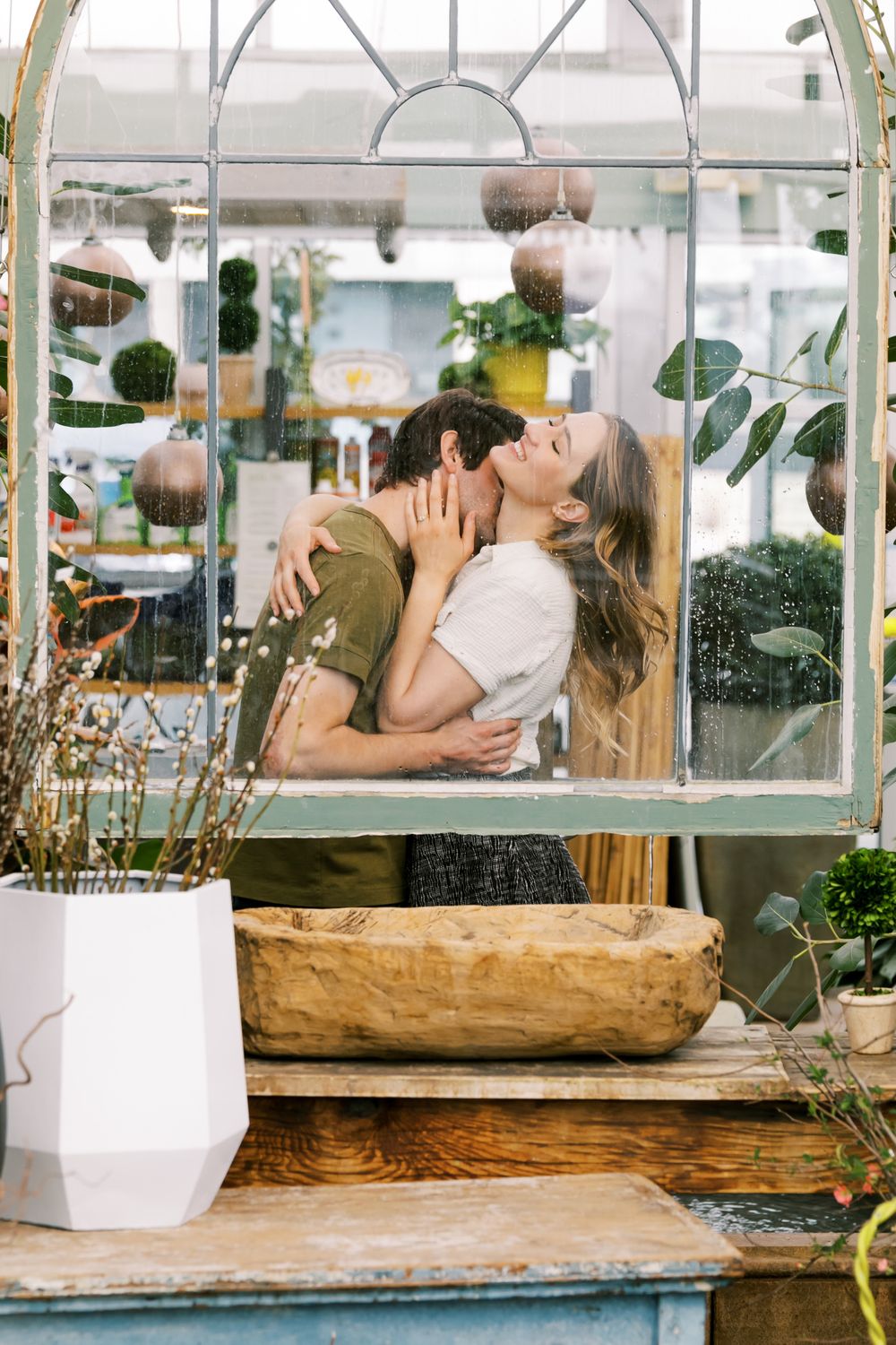 A romantic moment captured through a greenhouse window with plants and decor.