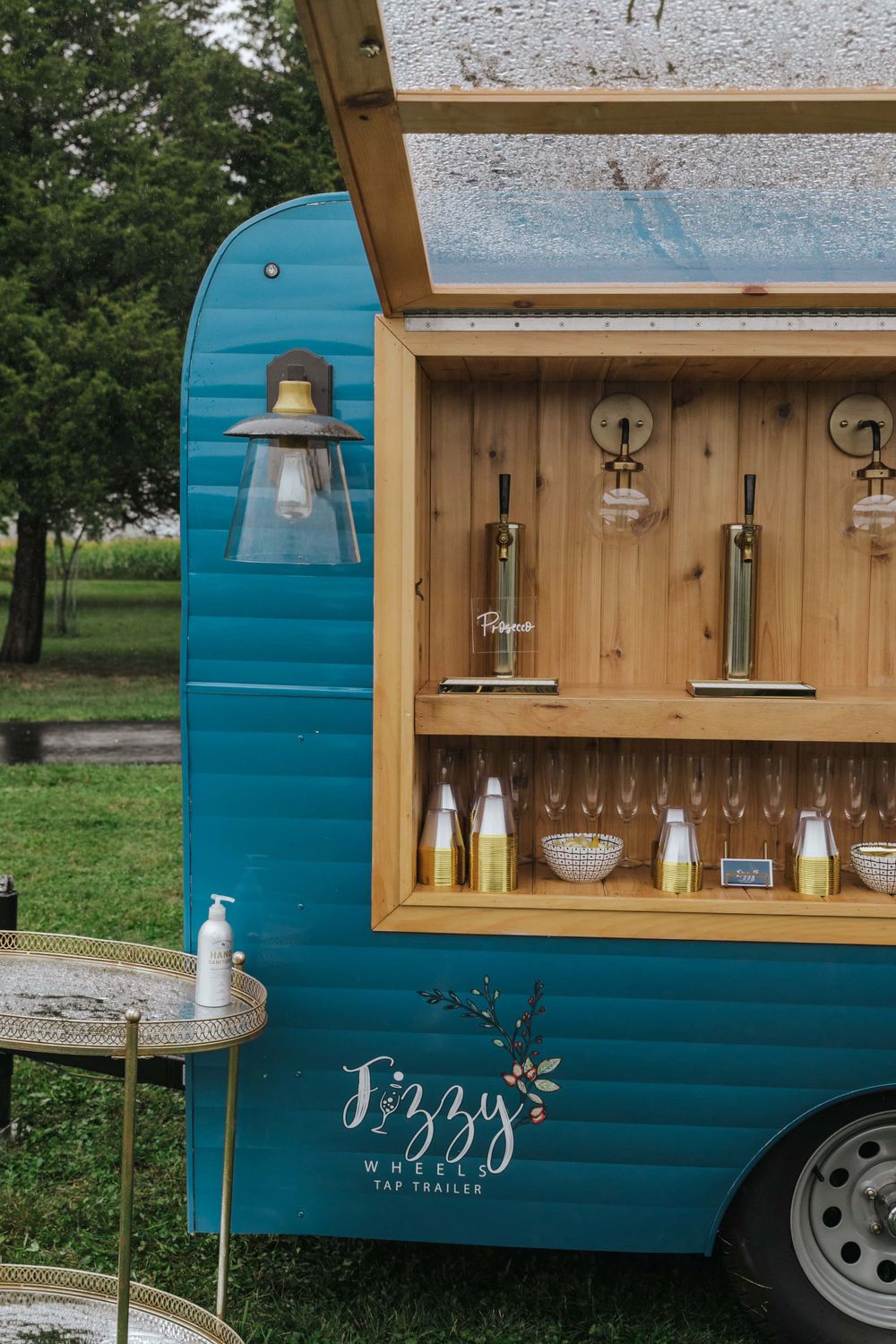A turquoise vintage mobile bar cart with wooden interior shelving displays glass bottles and cocktail supplies.