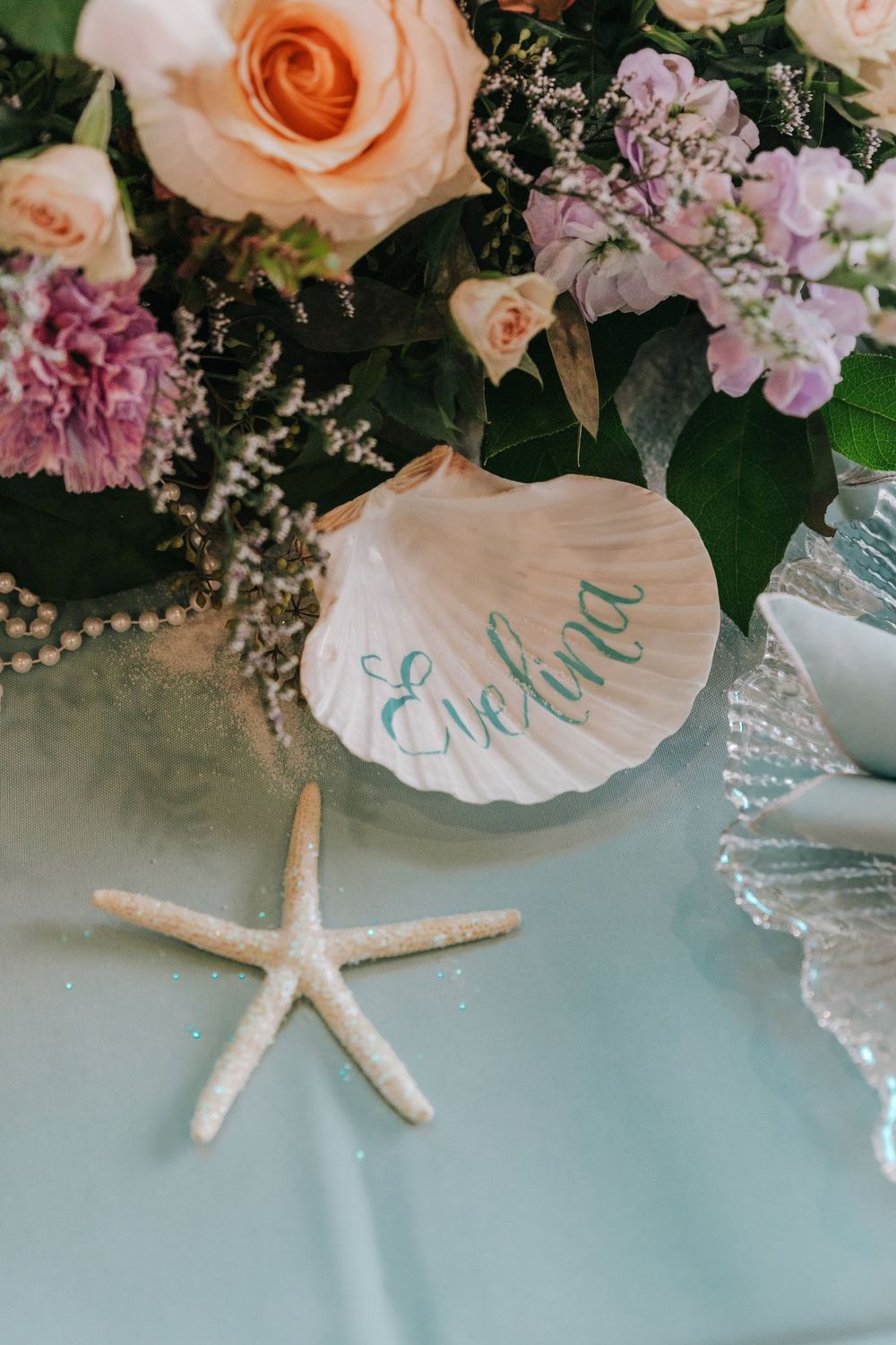A beach-themed wedding decor featuring a seashell with text, peach roses, pink carnations, and a white starfish on blue fabric.