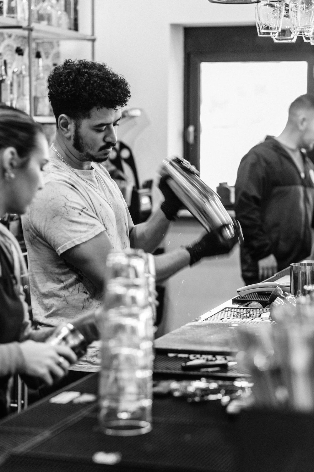 A bartender in a gray t-shirt working behind a crowded bar counter shown in black and white photography.