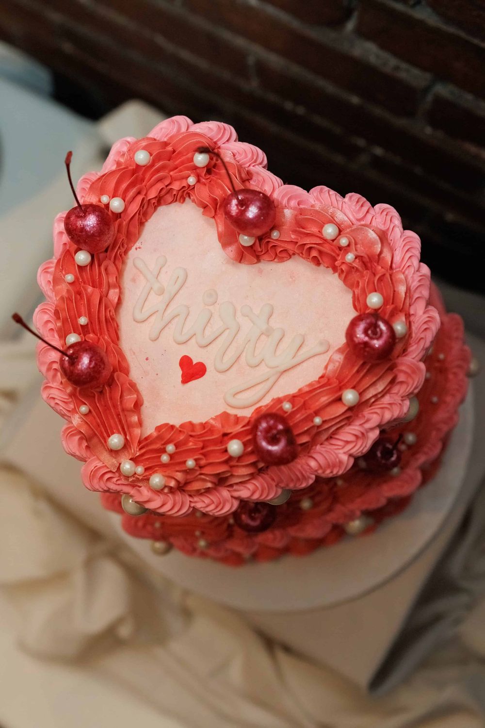 A heart-shaped pink cake decorated with red cherries, white pearls, and delicate piping details.