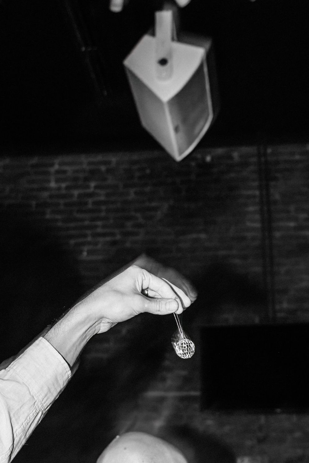 A black and white photo shows a hand dangling a disco ball beneath a light fixture against a brick wall.