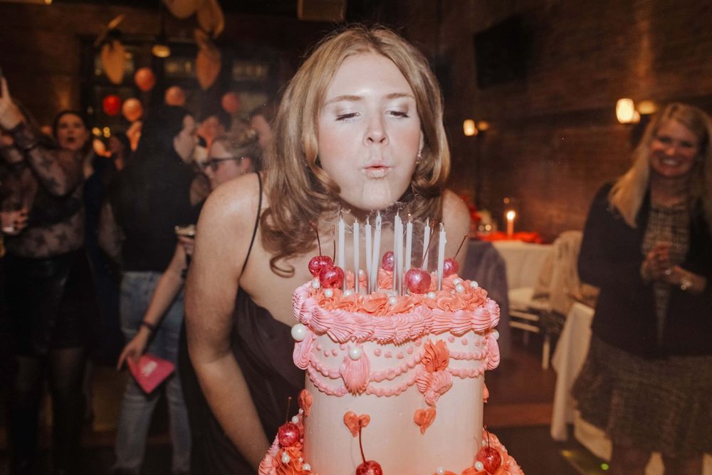 A person blowing out candles on a pink birthday cake at a dimly lit celebration party.