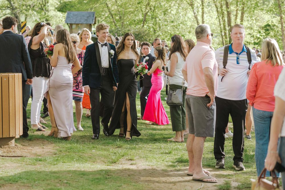 Wedding guests in formal attire gather outdoors on a sunny day during a celebration in a garden setting.
