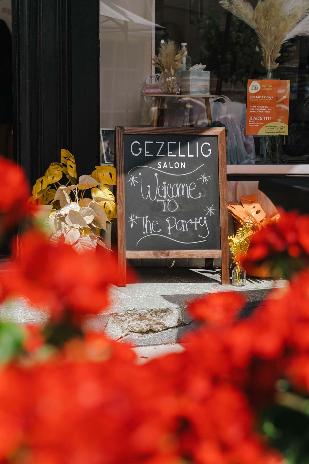 Chalkboard sign reading 'Gezellig Salon Welcome to the Party' framed by red flowers in the foreground.