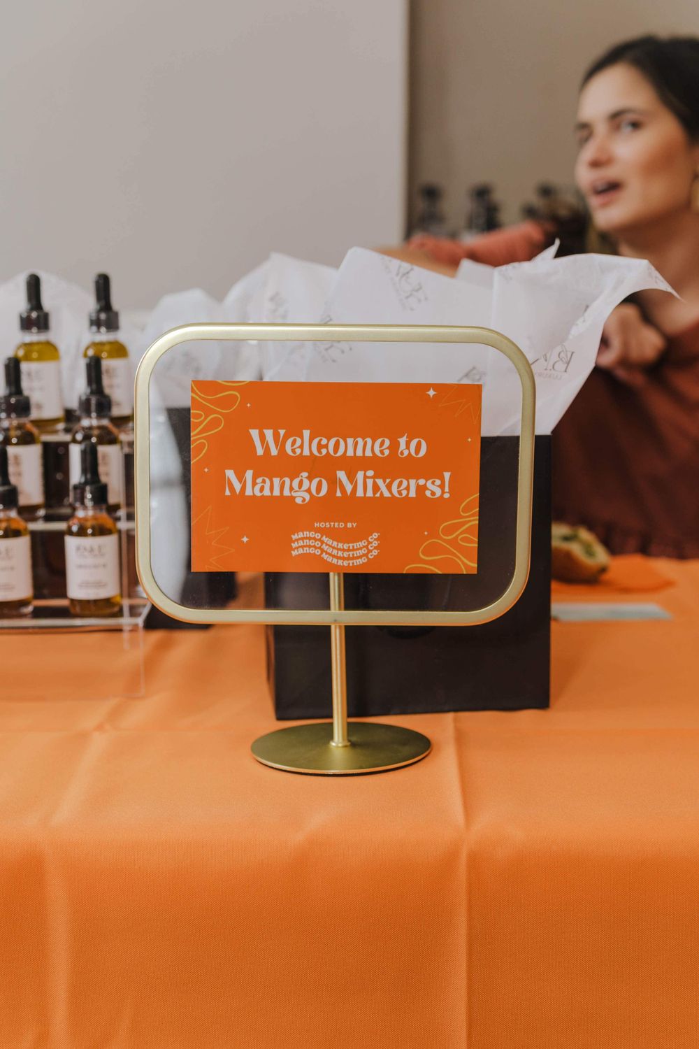A small orange sign reading 'Welcome to Mango Mixers!' sits on a table with amber bottles and an orange tablecloth.