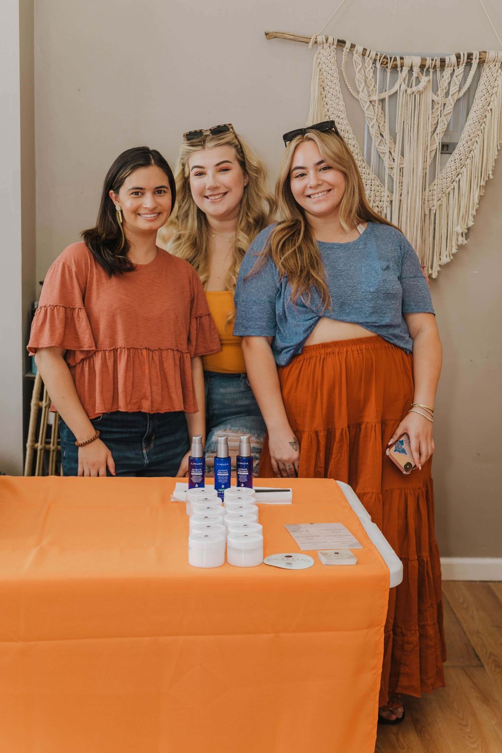 Three friends stand together at an event table with drinks and supplies against a white wall with macrame decor.
