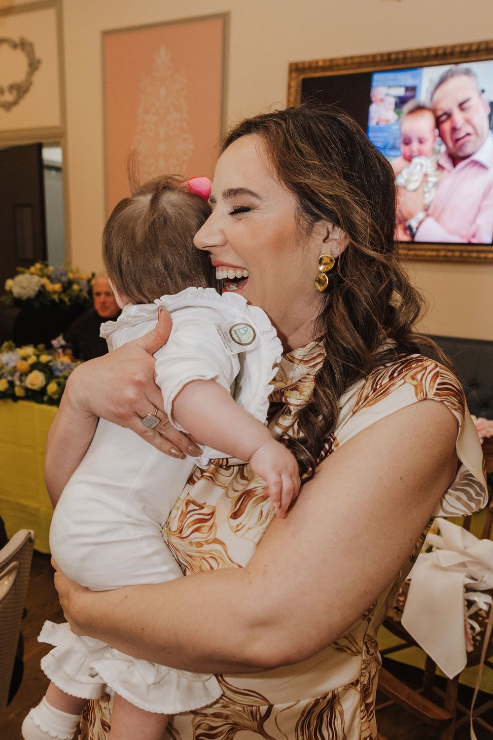 A joyful moment captured as someone in a floral dress laughs while holding a baby wearing white ruffled clothing.