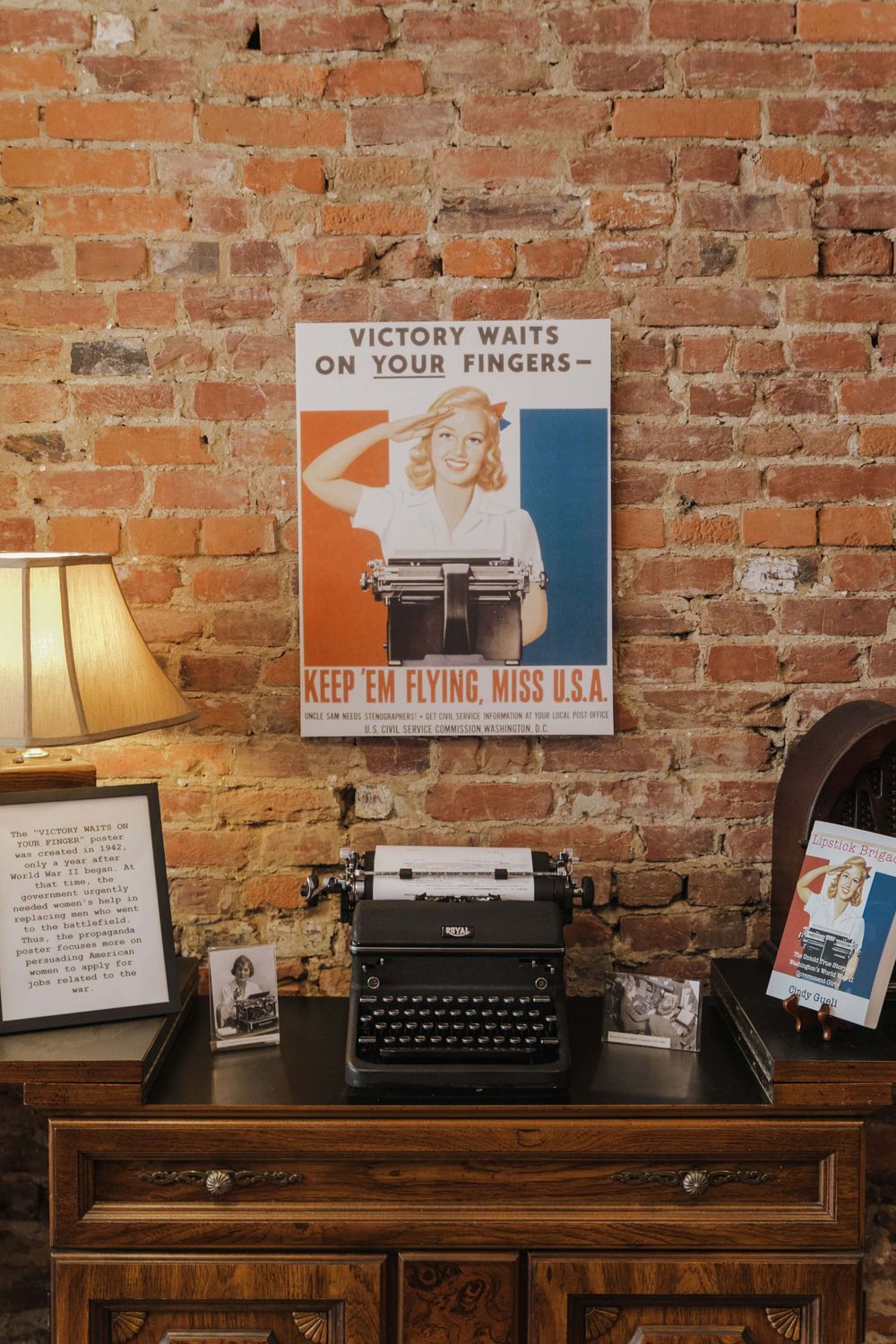 Vintage typewriter and lamp on wooden desk with WWII-era victory poster on exposed brick wall background.