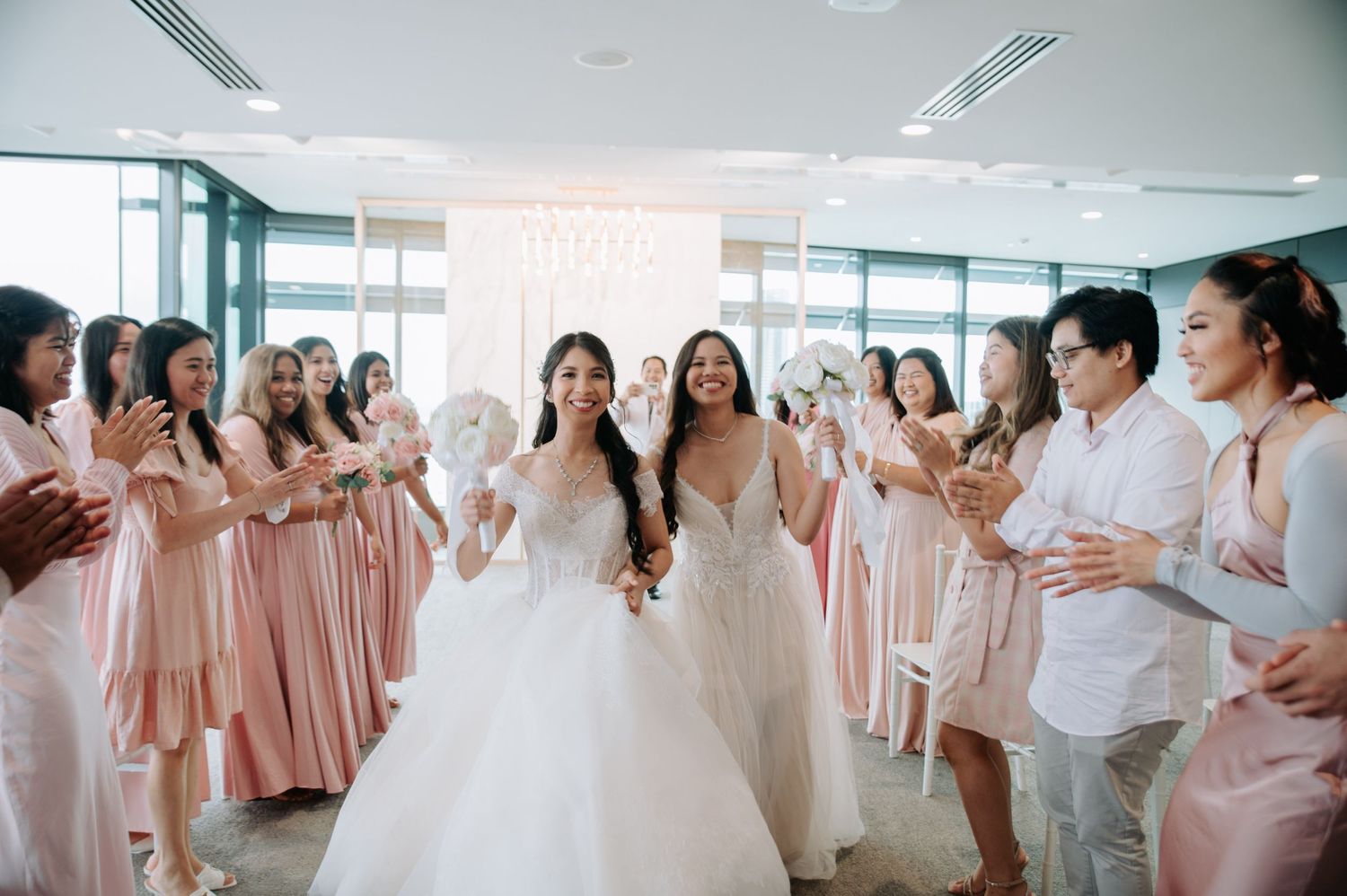 Wedding party in matching pink bridesmaid dresses celebrating with two brides in white gowns at a bright indoor venue.