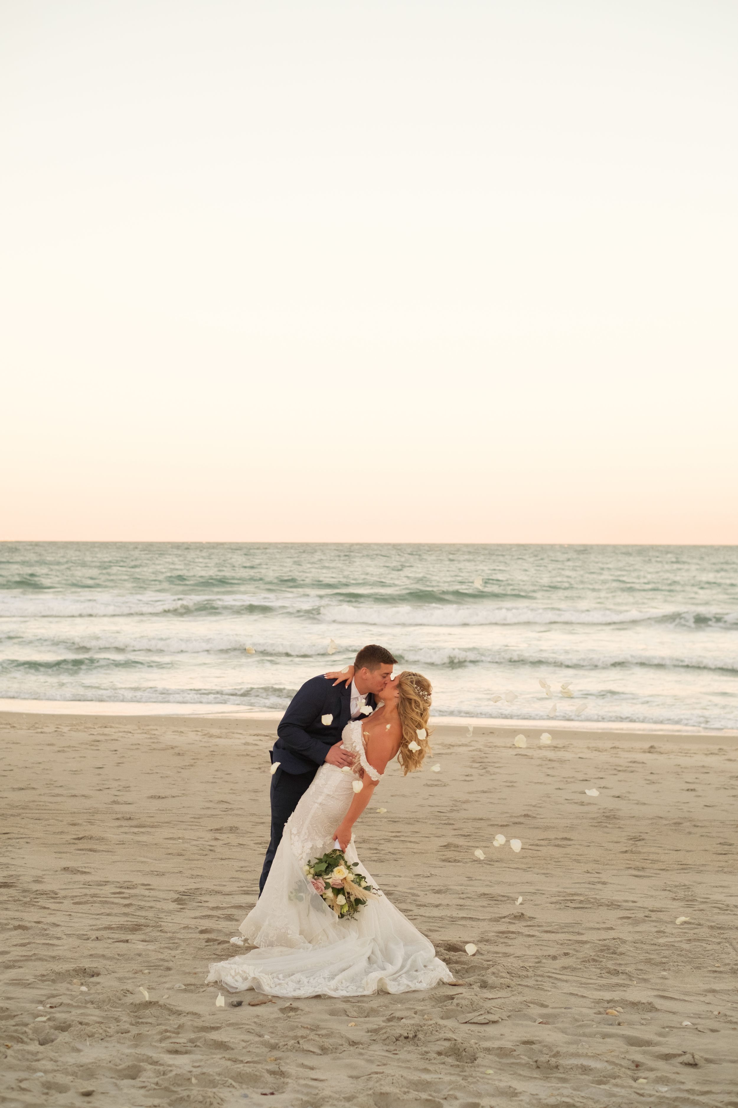 Flower petals fly past the bride and groom while they are kissing on the Florida beach with the sunset in the background