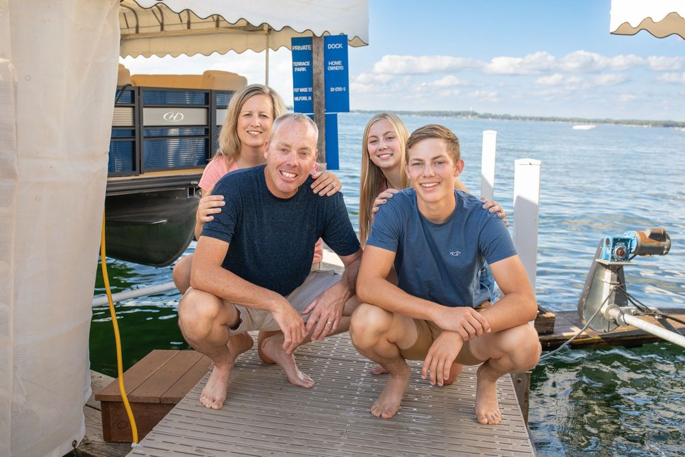 A family photo session of a family enjoying a summer day on a dock by West Okoboji Lake with boats visible in the background.