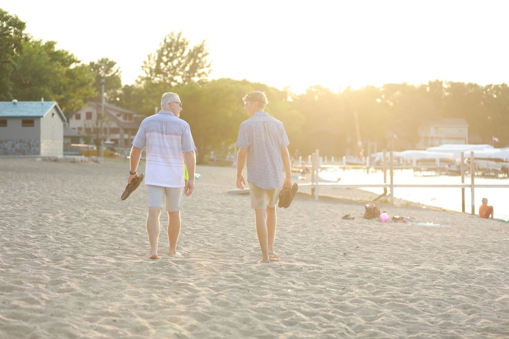 Father and son walking along a sandy beach in Okoboji, Iowa at sunset with golden sunlight streaming through the trees in the background.
