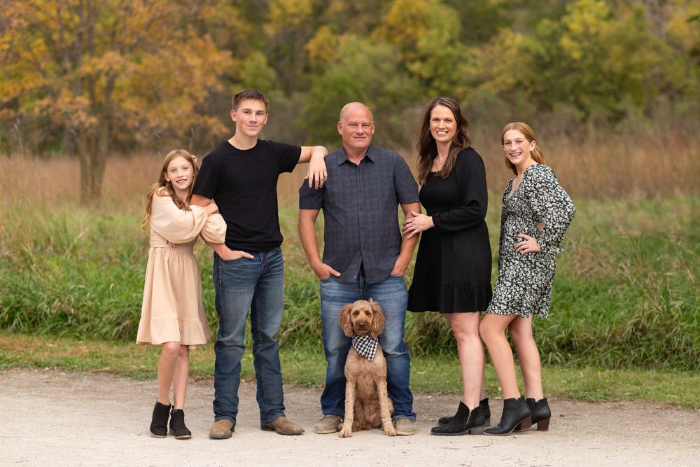 A family and their dog pose together outdoors on a path during autumn with fall foliage in the background at Adam's Nature Preserve in SD.