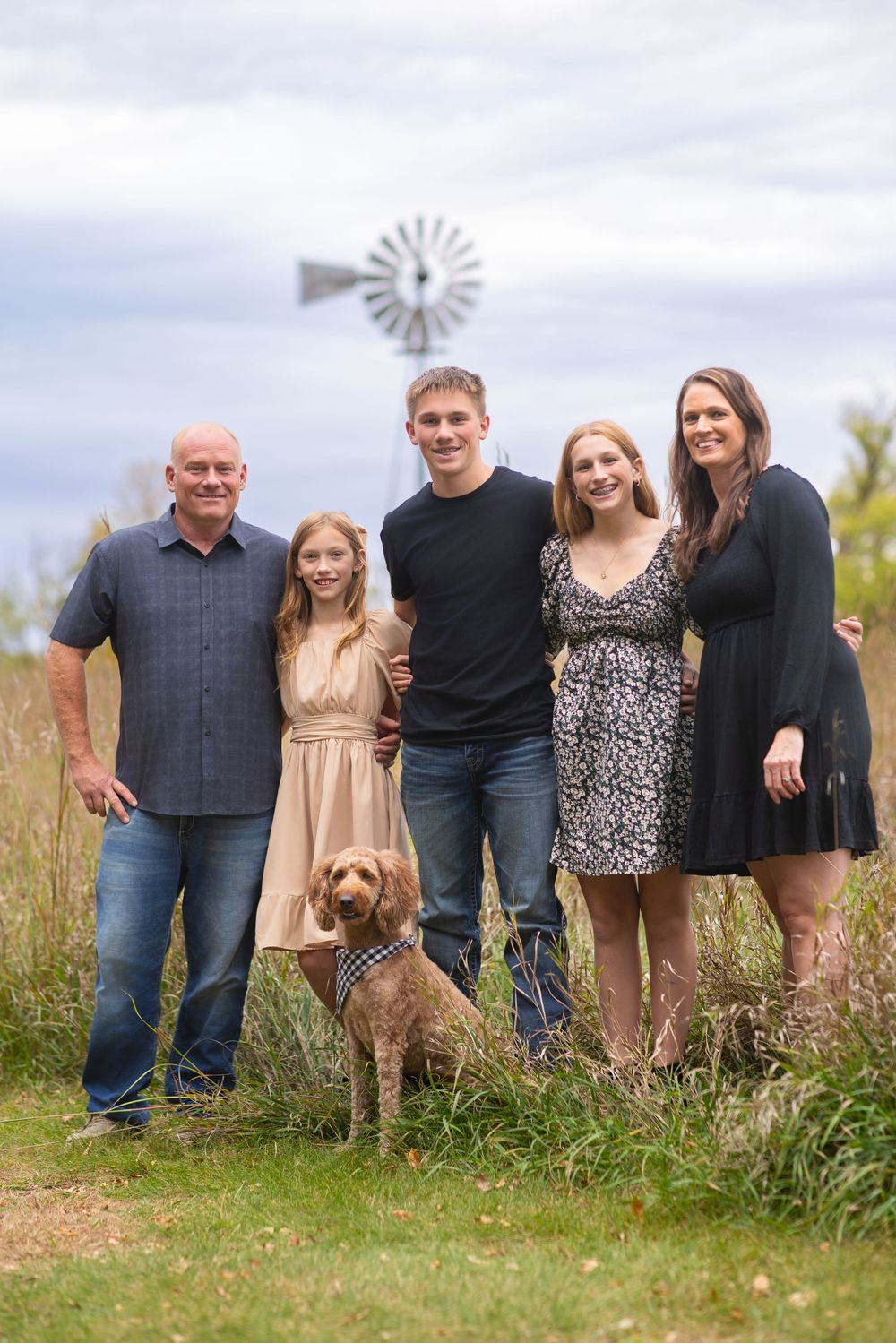 A family stands together in a field with their family dog while a windmill spins in the background at Adam's Nature Preserve.