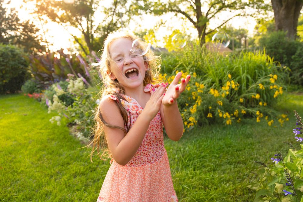 Joyful child wearing pink floral dress laughs as she pops bubbles in sunny garden at Latham Park with yellow flowers and green grass.