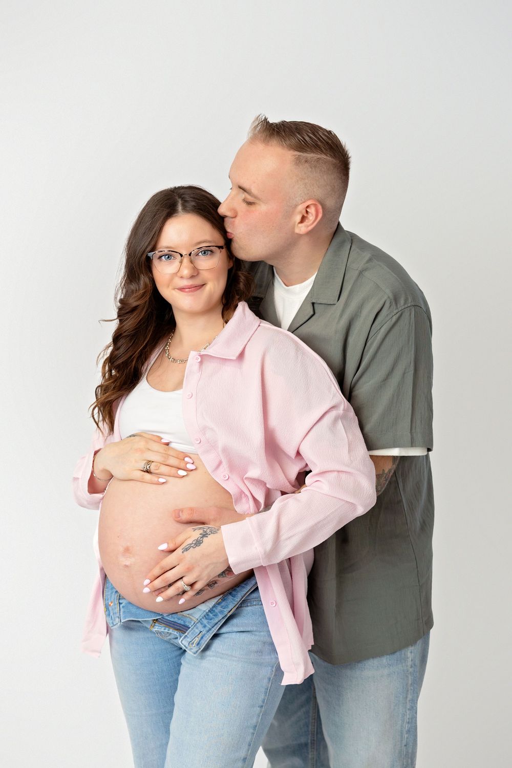 Couple in casual attire sharing intimate moment against white background in maternity photo series.