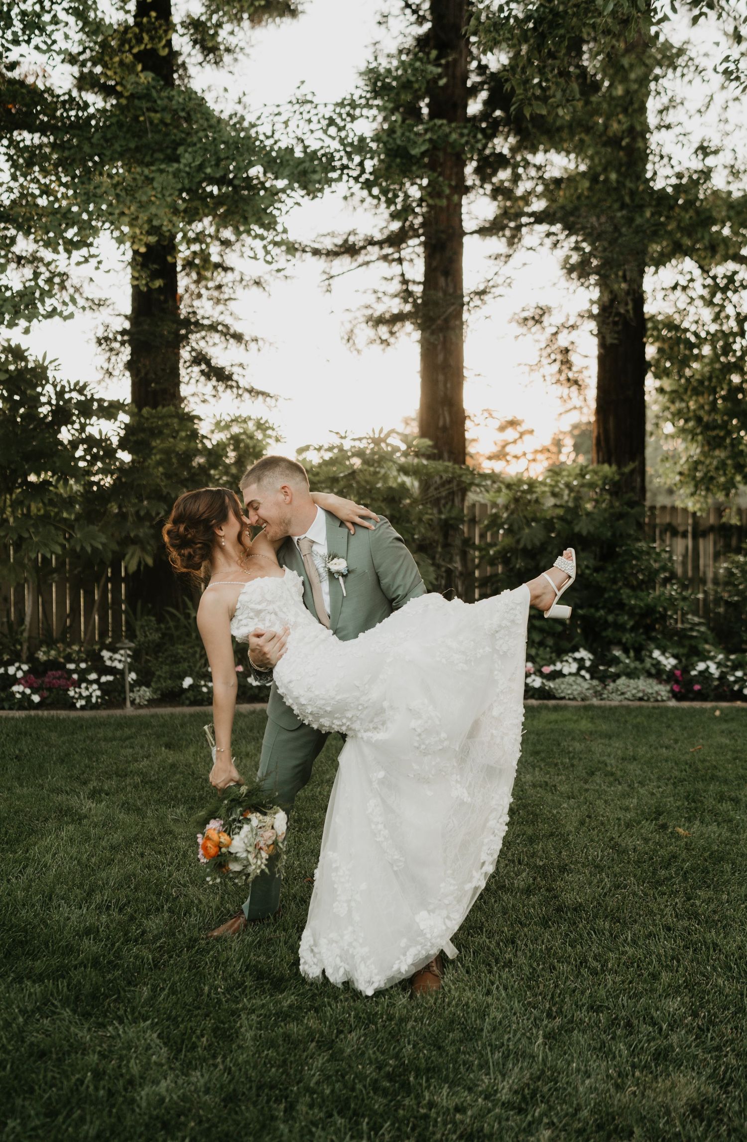 Romantic outdoor wedding photo with dramatic dip kiss on a garden lawn at sunset.