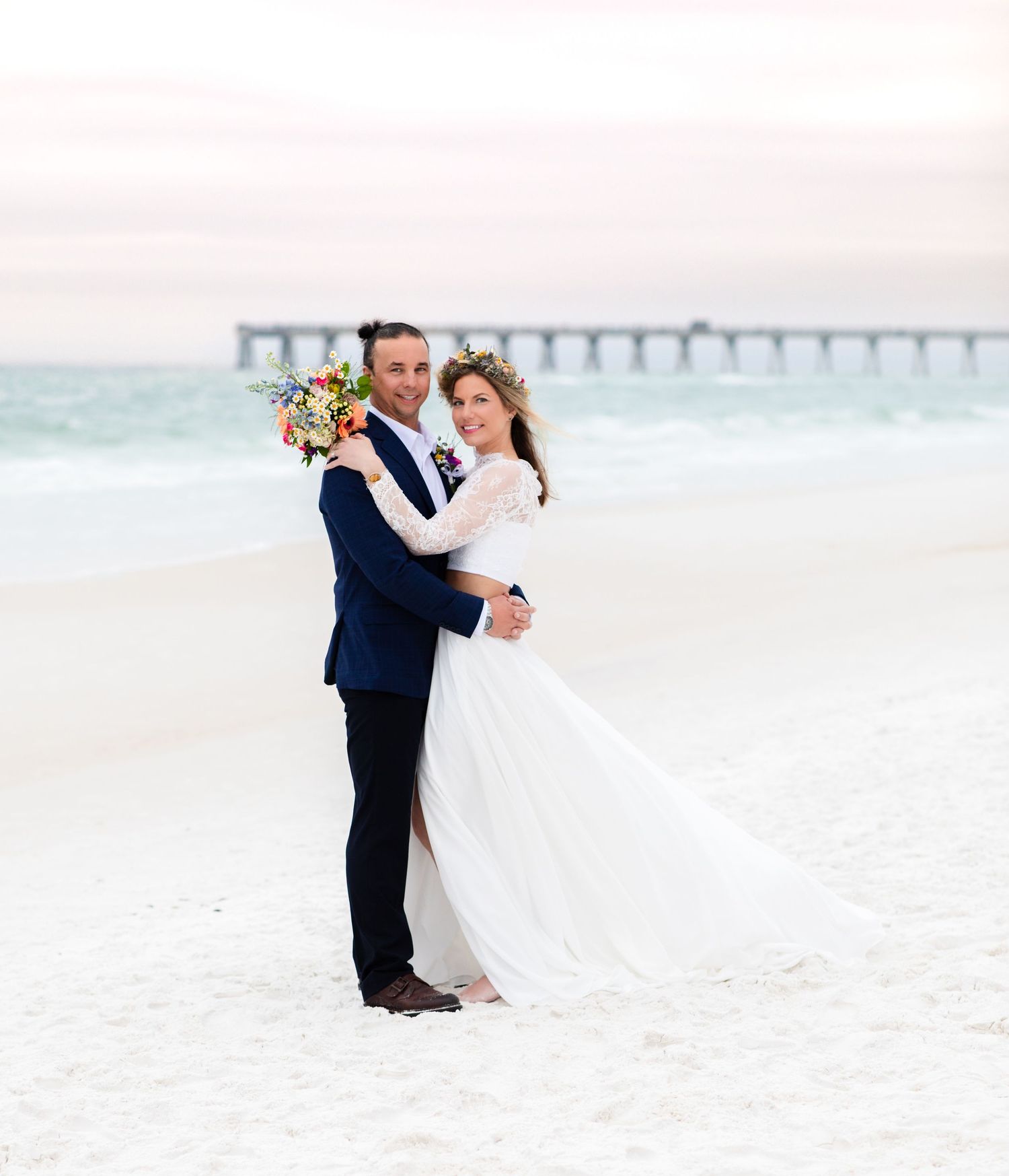 A newlywed couple poses on a beach with a pier in the background.