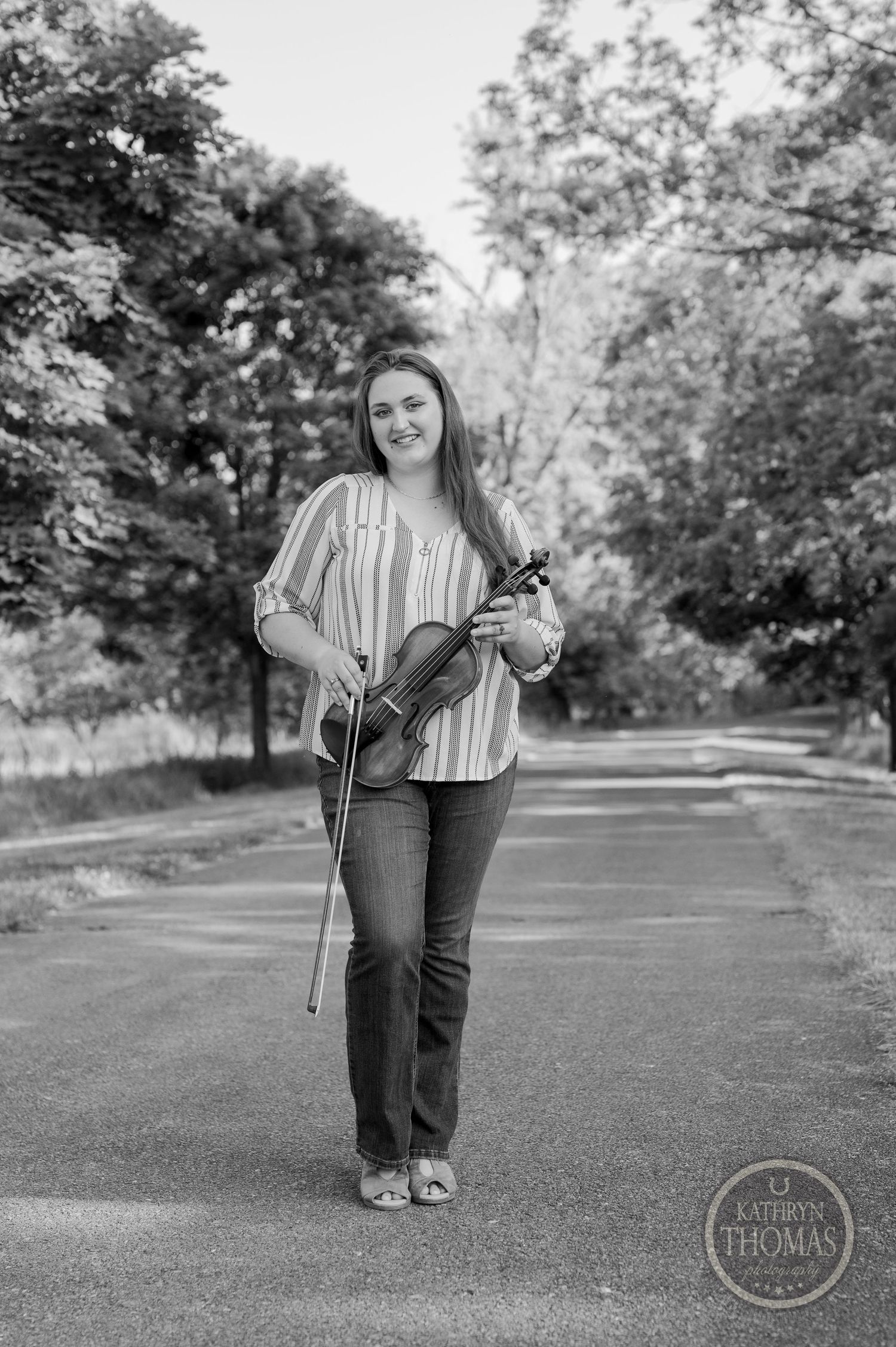 Black and white portrait of violinist standing on pathway surrounded by trees.