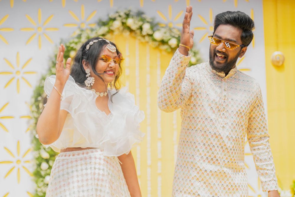 Couple in white traditional outfits celebrating against a yellow backdrop with floral decorations.