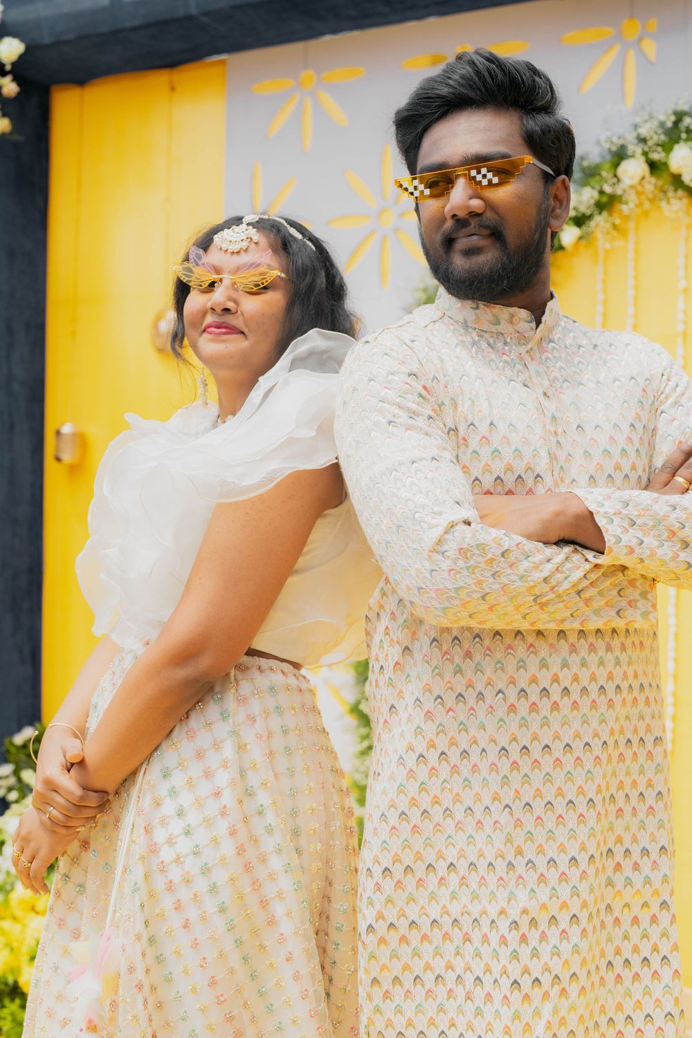 Stylish couple in coordinated white outfits posing against a yellow wall with floral decor.