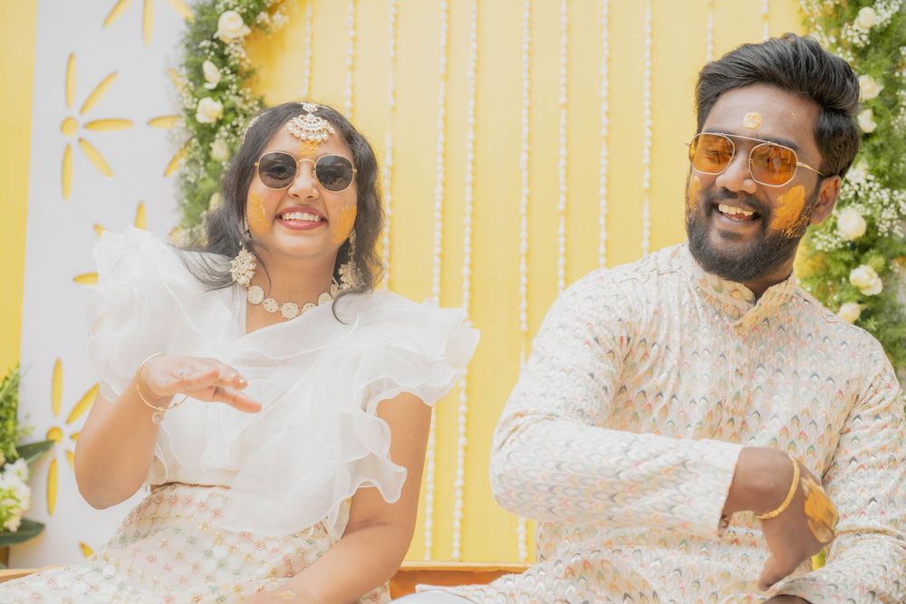 Two people wearing white outfits sit together smiling against a yellow background with floral decorations.