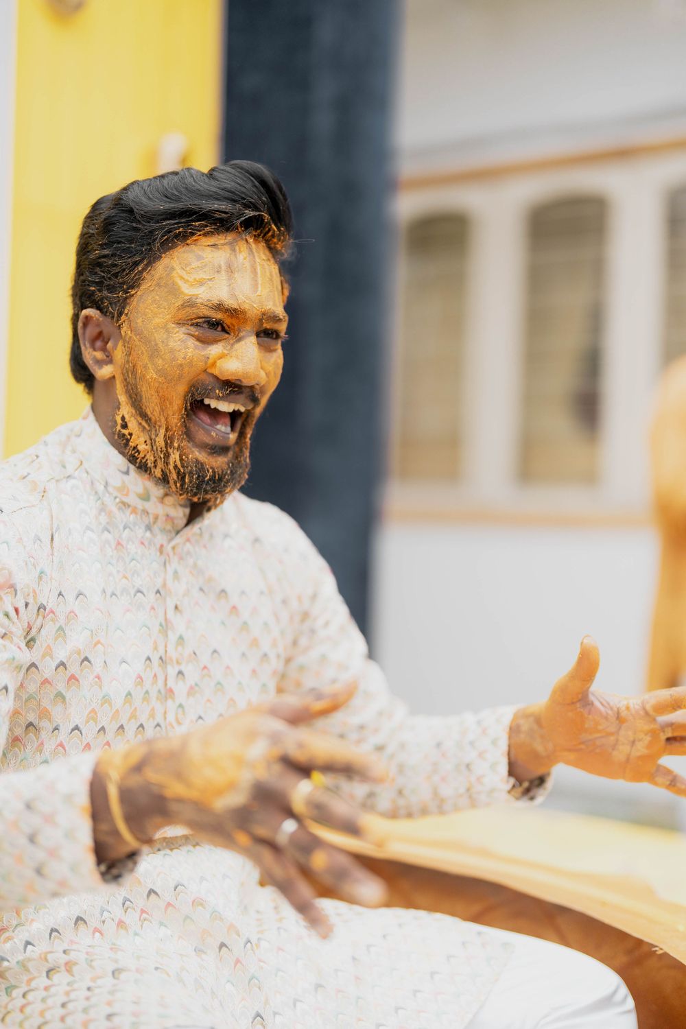 Person in white traditional attire laughing joyfully with turmeric powder on face.