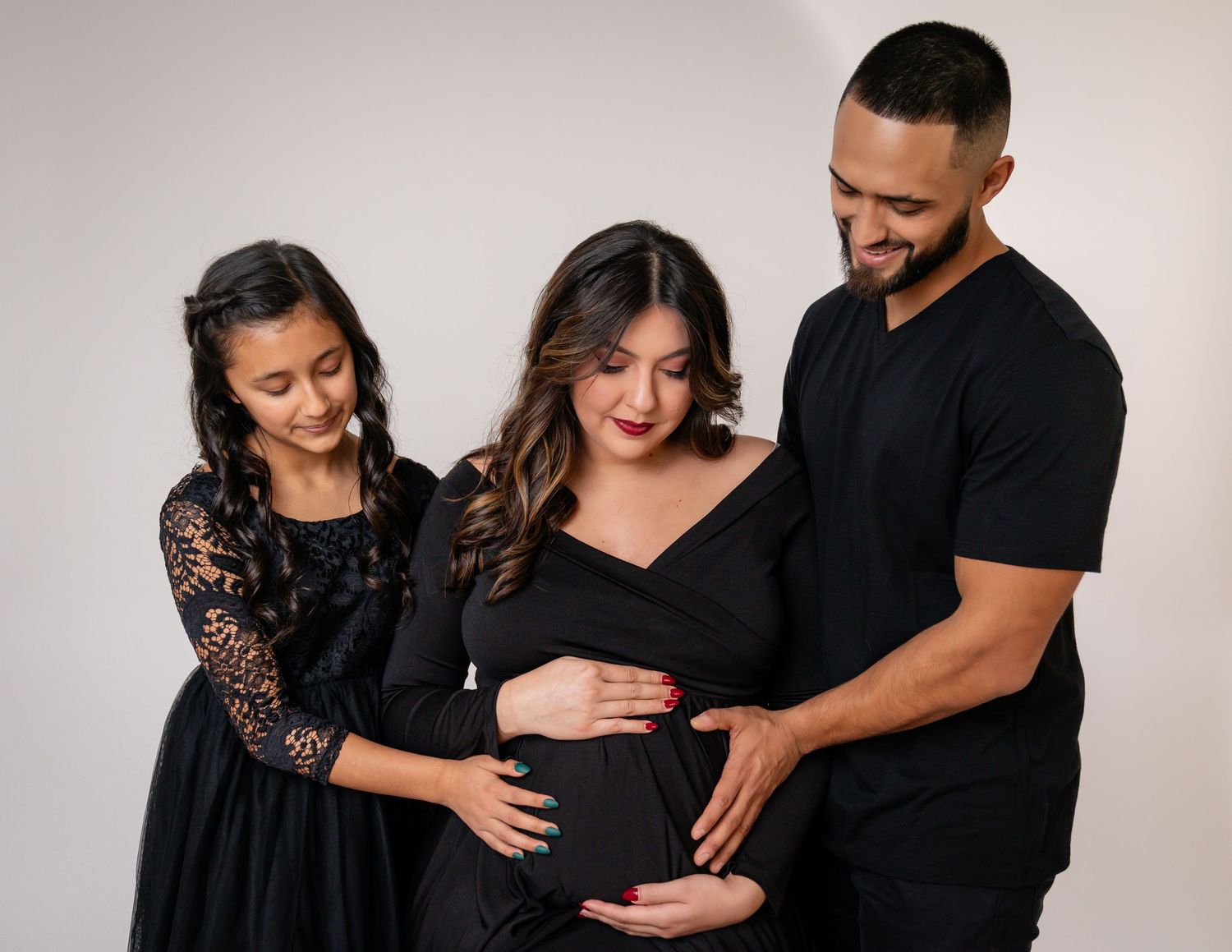 Family maternity photo session featuring three people in black outfits against white backdrop.