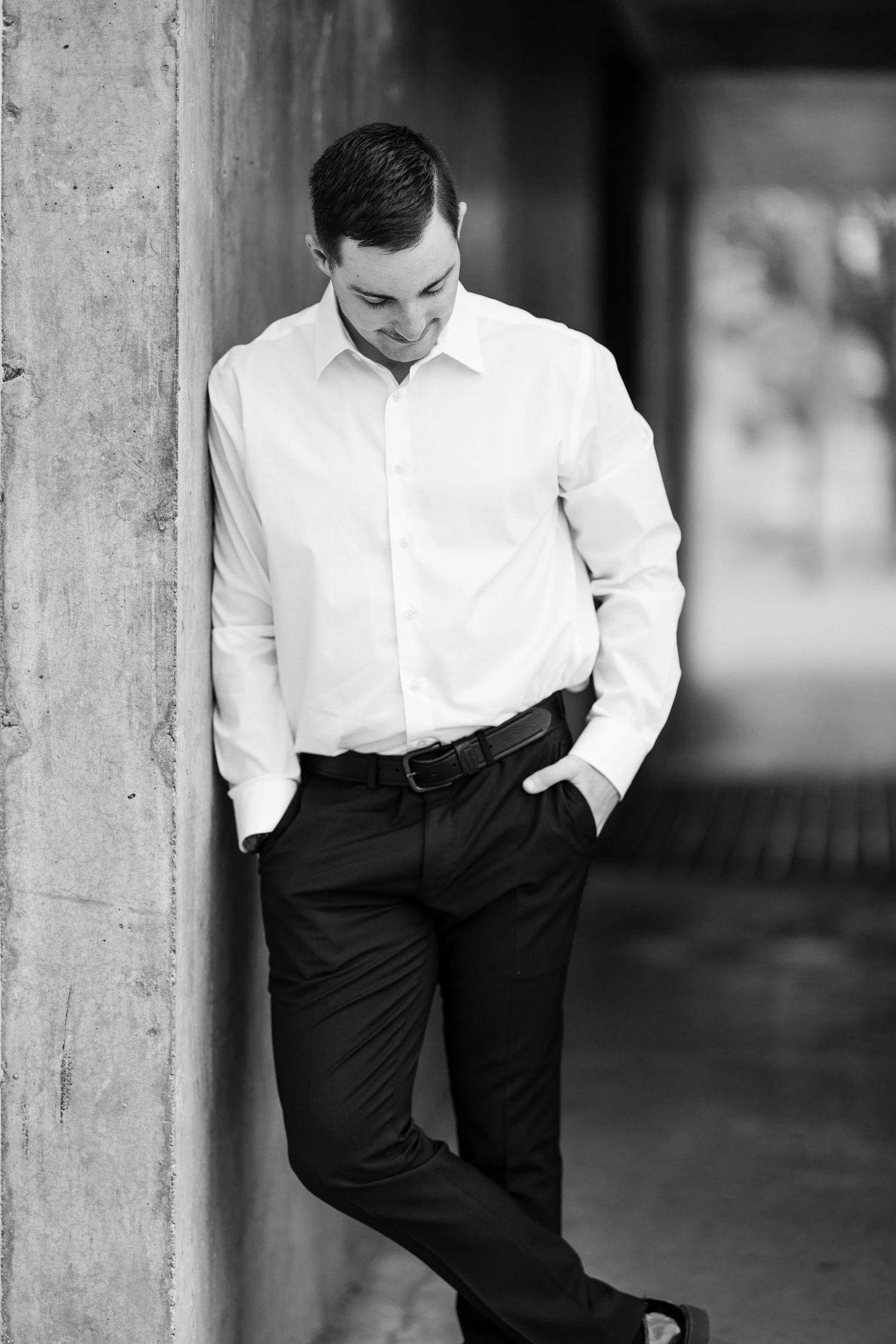 Professional black and white portrait of a person in business attire leaning against a wall.