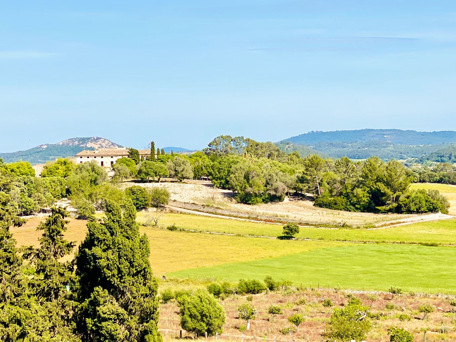 Rolling green countryside with mountains and Mediterranean architecture in the distance under blue skies.