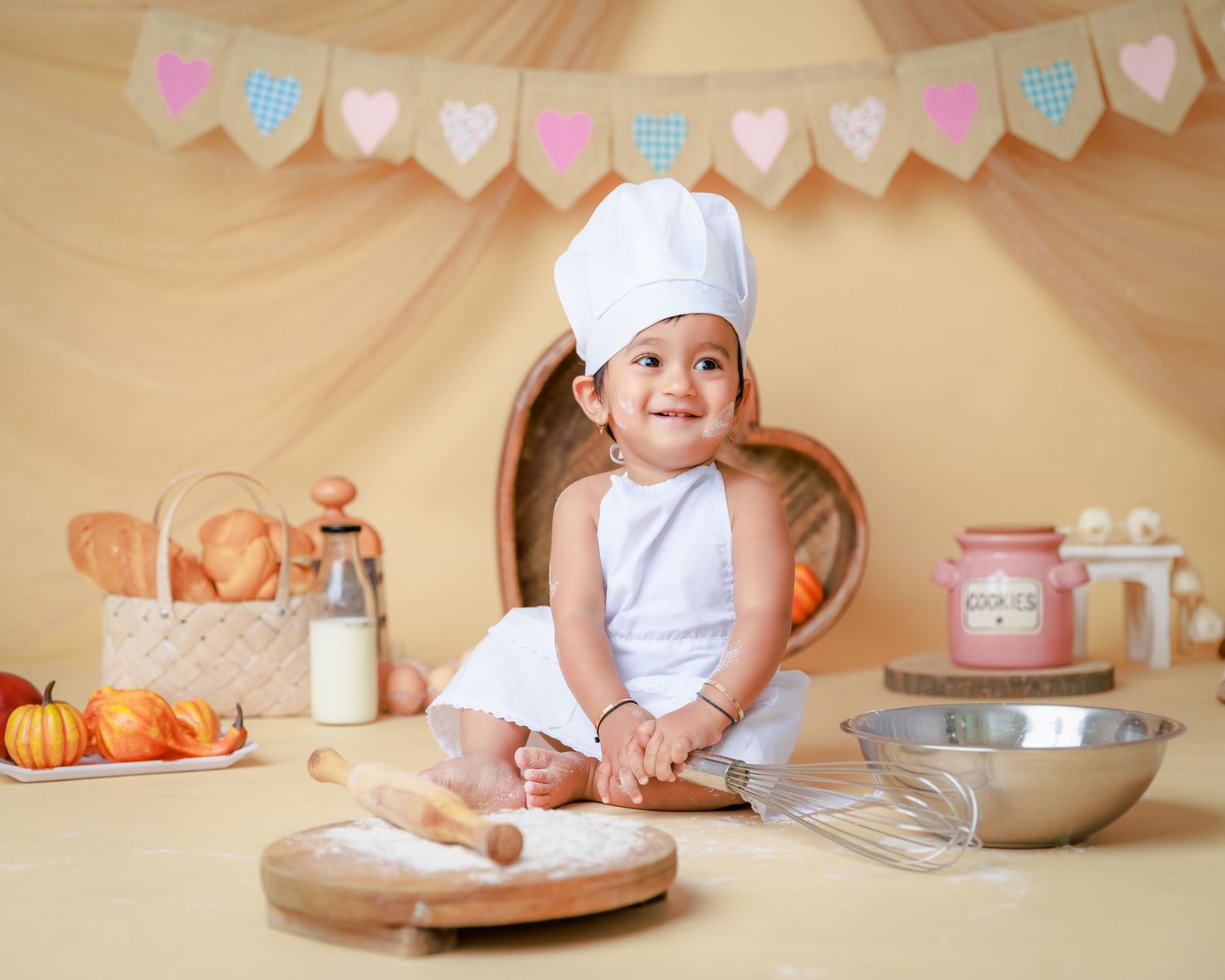 Baby in chef outfit sits with baking ingredients and utensils under party banner.