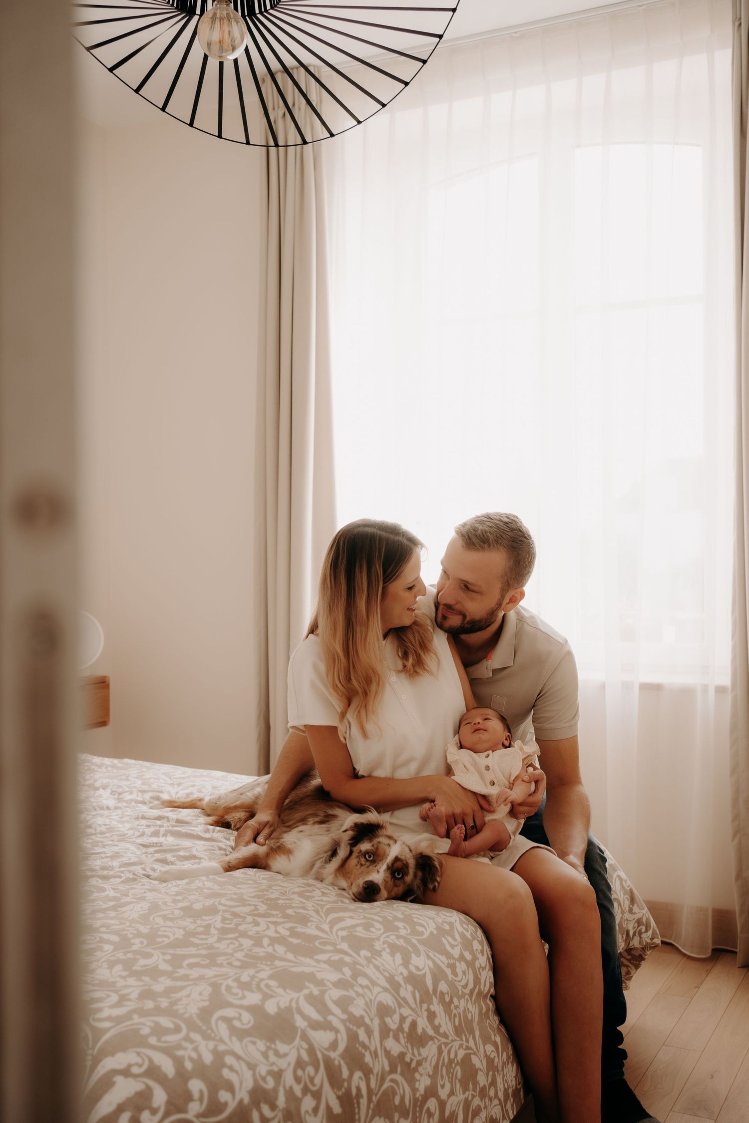 Family bonding with newborn on white bedding in a bright, minimalist bedroom.