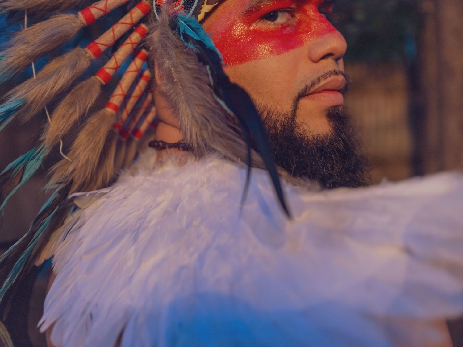 Native American man in traditional feathered headdress with red face paint and white feathers in dramatic lighting.