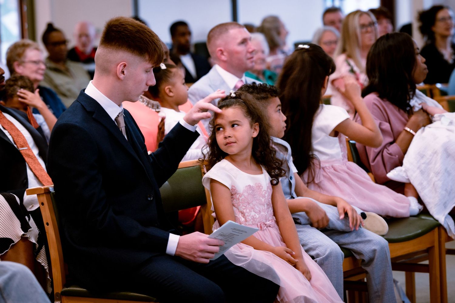 Parent and child in formal attire share a tender moment during a ceremony while seated among other attendees.