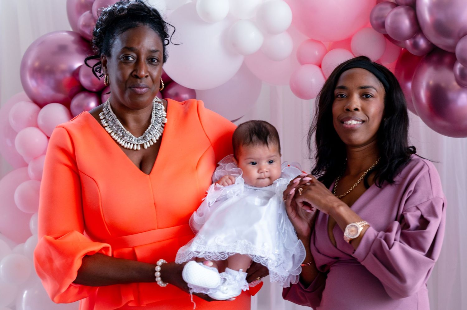 Three generations pose together against a pink balloon backdrop at a special celebration.