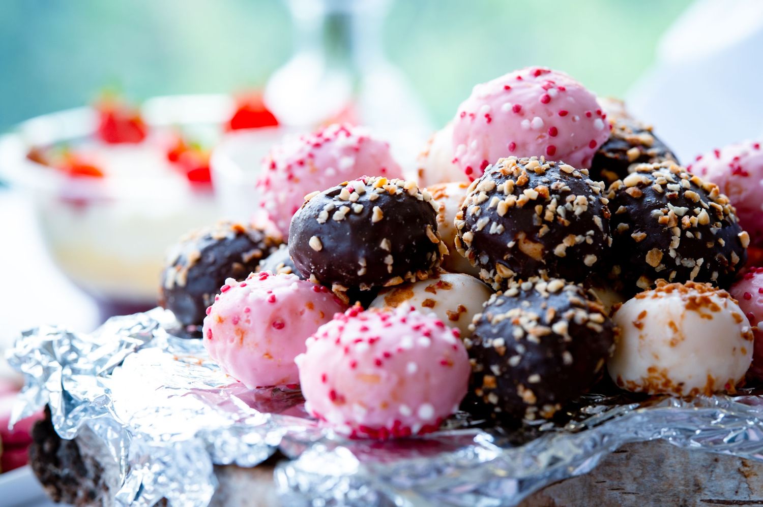 Chocolate and pink cake pops decorated with sprinkles and sesame seeds displayed on silver foil with blurred background.