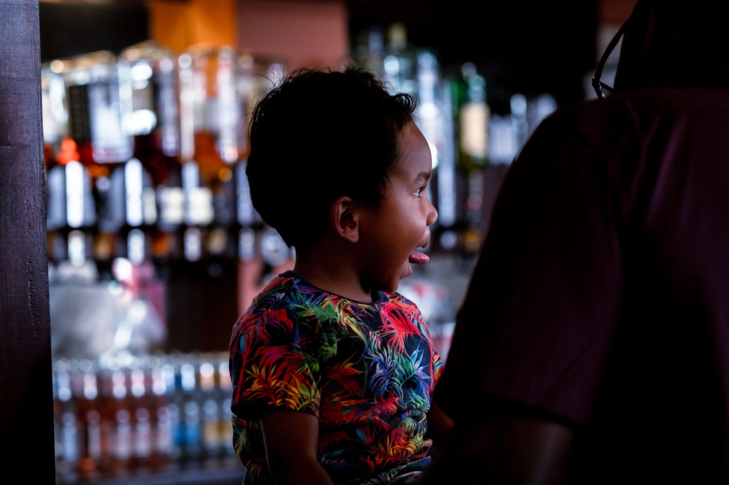 A child in a floral shirt stands at the edge of a blurred bar with bottles in the background.